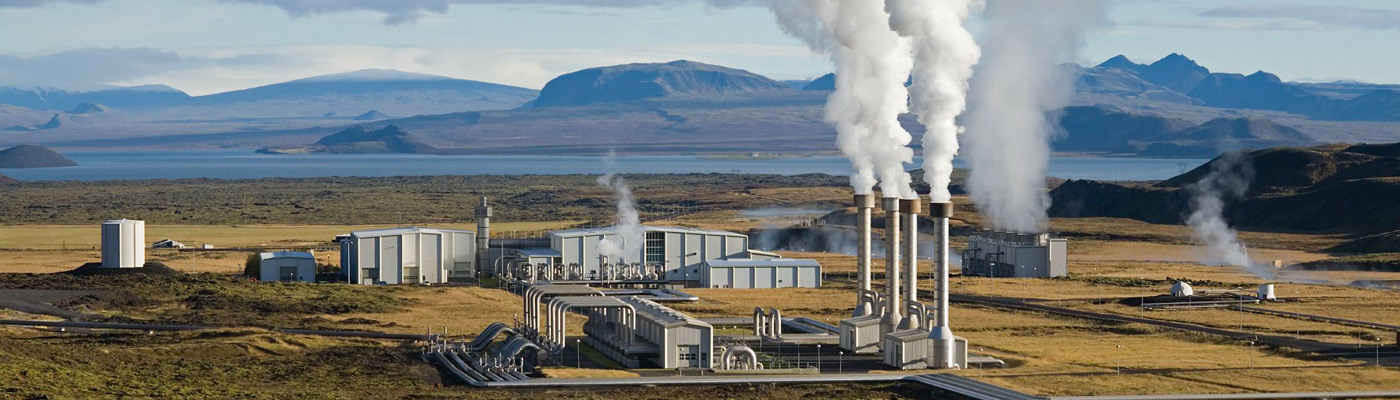 A power station with smoke billowing from chimneys, near a sea and mountain landscape