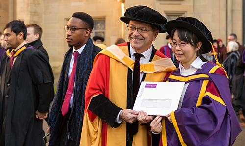 A female research graduate in her gown receiving a certificate from her supervisor