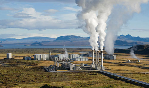 A power station with smoke billowing from chimneys, near a sea and mountain landscape